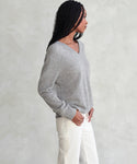A woman with long braided hair wears the Flynn Cashmere Sweater in light gray and off-white pants, standing sideways against a textured light background.