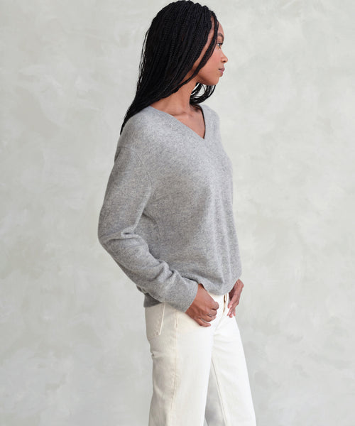 A woman with long braided hair wears the Flynn Cashmere Sweater in light gray and off-white pants, standing sideways against a textured light background.
