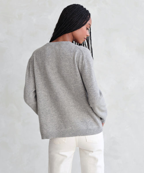 A person with long braids stands with their back to the camera, wearing the Flynn Cashmere Sweater in light gray and off-white pants against a light, textured background.
