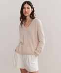 A woman with wavy brown hair wears the Flynn Cashmere Sweater, a relaxed fit knit V-neck, paired with cream-colored shorts, standing against a plain light background with her hands in her pockets.