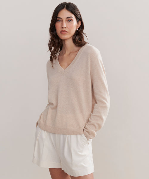 A woman with wavy brown hair wears the Flynn Cashmere Sweater, a relaxed fit knit V-neck, paired with cream-colored shorts, standing against a plain light background with her hands in her pockets.
