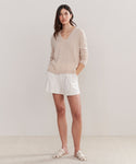 A woman stands against a plain light background wearing the Flynn Cashmere Sweater in light beige, styled with white shorts and white sandals. Her hands are in her pockets as she looks at the camera with a neutral expression.