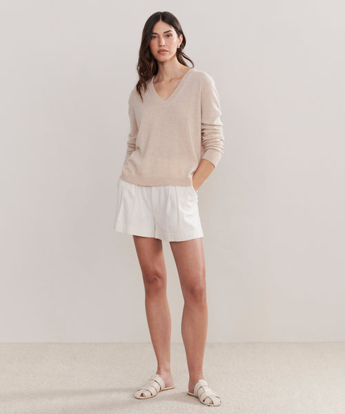A woman stands against a plain light background wearing the Flynn Cashmere Sweater in light beige, styled with white shorts and white sandals. Her hands are in her pockets as she looks at the camera with a neutral expression.