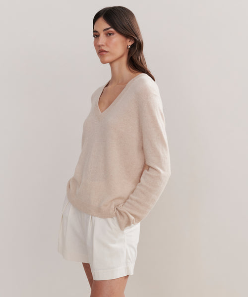 A woman stands against a light background, wearing the Flynn Cashmere Sweater in beige and white shorts. Her hands are in her pockets, and she has a calm, neutral expression.