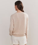 A woman with long brown hair is seen from behind, wearing the Flynn Cashmere Sweater in a relaxed fit and light beige, paired with off-white shorts, standing against a plain light background.
