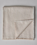 The Frayed Linen Tablecloth in beige and white natural stripes is folded with frayed edges, shown on a light grey surface. One corner is flipped to display the underside.