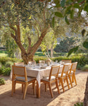 A wooden outdoor dining table with eight chairs is set under a leafy tree in a sunny garden, featuring plates, glasses, and the Frayed Linen Tablecloth with natural stripes and frayed edges, surrounded by greenery and mountain views.