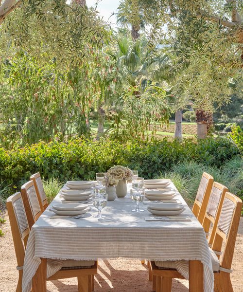A wooden outdoor dining table covered with the Frayed Linen Tablecloth is styled with plates, bowls, glasses, and a floral centerpiece. Six chairs surround the setup amid lush garden greenery and trees in the background.