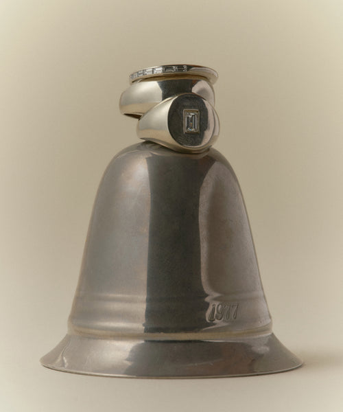 A shiny silver bell engraved with “1977” holds two class rings, one upright and one angled. A Gemma Baguette Signet Ring adds sparkle, set against a soft neutral gradient background.