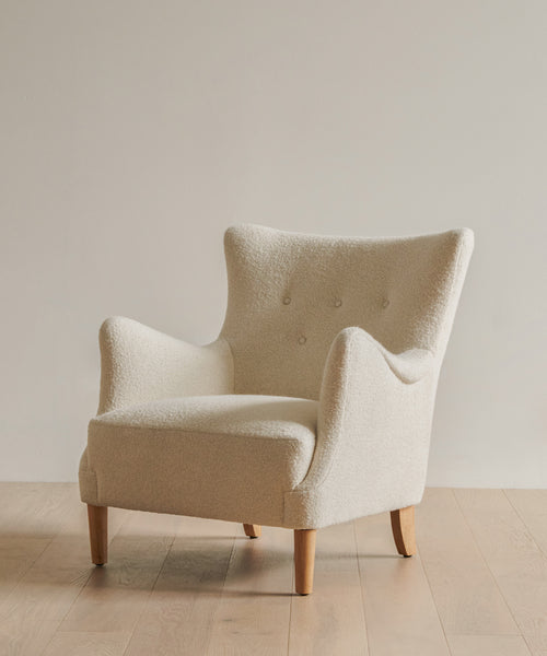 A modern Greenwich Chair with cream Mini Boucle upholstery, wooden legs, and a curved backrest, handmade in the USA. Pictured on a light wooden floor with a plain off-white wall background.