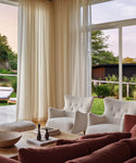 A cozy living room features cream-colored Greenwich Chairs and sheer white curtains, a wooden coffee table with decor, rust sofas, and large windows revealing a lush green garden and patio outside.
