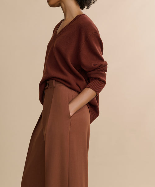 A person stands against a beige background wearing a loose brown V-neck sweater with sleeves pushed up and high-waisted Greta Trouser, which has a relaxed drape. One hand is in their pocket, and their face is not visible.