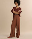 A woman stands with arms crossed, wearing high-waisted Greta Trousers with a relaxed drape and a rust sweater, styled with matching shoes and belt, set against a beige backdrop.