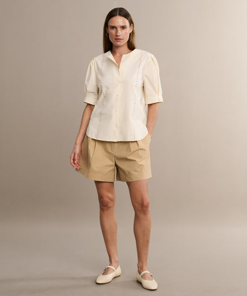 A woman stands against a beige background wearing the Harper Blouse in Italian cotton poplin, paired with tan shorts and cream flat shoes. She gazes at the camera with a neutral expression and relaxed posture.
