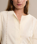 A woman wears the Harper Blouse, an Italian cotton poplin button-up with embroidered eyelets along the front seams, shown in cream from shoulders to mid-torso.
