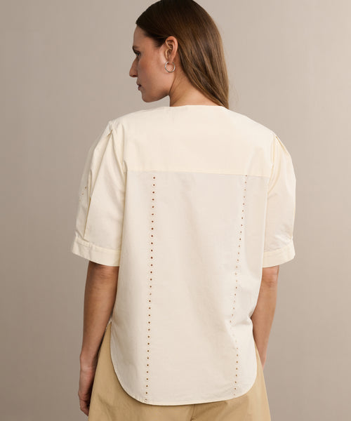 A woman with straight brown hair models the Harper Blouse, a cream Italian cotton poplin top with short sleeves and embroidered eyelets at the back seams. She stands with her back to the camera against a neutral background.