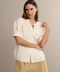 A woman in the Harper Blouse, a cream Italian cotton poplin top with embroidered eyelets, pairs it with beige shorts and stands against a beige background, gazing to the side and touching her collar.
