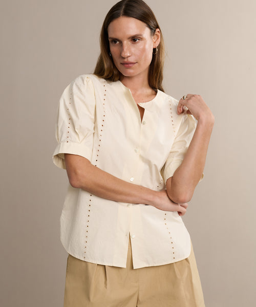 A woman in the Harper Blouse, a cream Italian cotton poplin top with embroidered eyelets, pairs it with beige shorts and stands against a beige background, gazing to the side and touching her collar.