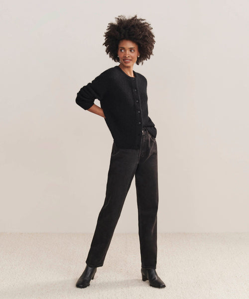 A person with curly hair stands against a plain background, wearing the black Hayes Crewneck Cardigan, black jeans, and black ankle boots—one hand behind their back, the other in their pocket.