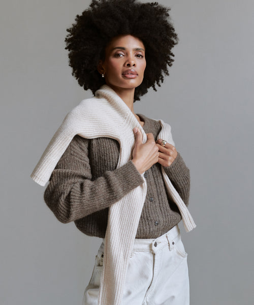 A woman with a voluminous afro wears the Hayes Crewneck Cardigan in brown and white pants, with a cream knit sweater draped over her shoulders, posing against a plain gray background.