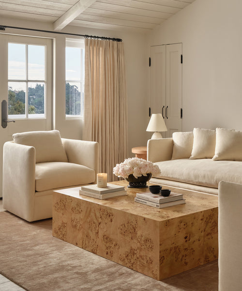 A modern living room with a cream sofa and the Dume Chair in beige hemp upholstery, a light wood coffee table, books, flowers, a candle, beige curtains, and soft natural light streaming through large windows.