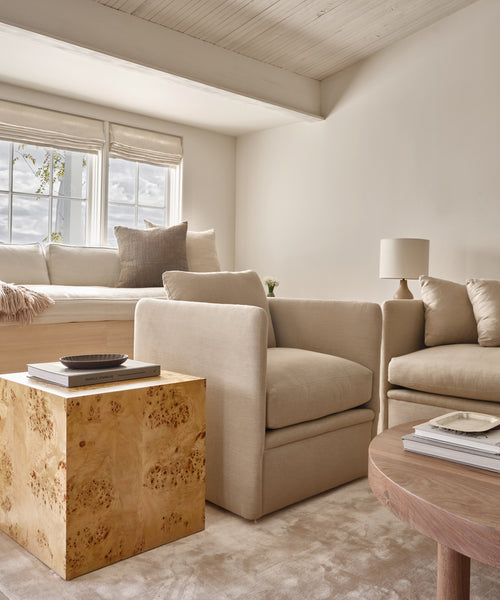 A bright, modern living room showcases sustainable pieces like the cream Dume Chair, a window seat with pillows, a beige sofa, and unique wood tables—all in soft neutrals with natural light streaming through the window.