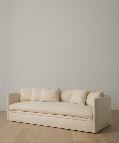 The Dume Sofa, featuring sustainable beige hemp upholstery and four matching cushions, sits on wooden flooring against a plain light-colored wall.