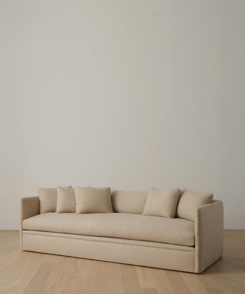 The Dume Sofa, a minimalist beige Hemp piece with five matching cushions and sustainable upholstery, sits on a light wooden floor against a plain off-white wall.