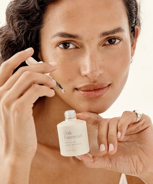 A woman with wavy hair holds a dropper and a bottle of The Hydration Heroes oil near her face, ready to apply it. She has natural makeup and a calm expression.