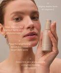 A woman holds a bottle of Daily Vitamin C Serum near her face. Text highlights its benefits: stable formula, anti-aging properties, brightening dark spots, firming skin, and supporting a radiant complexion.