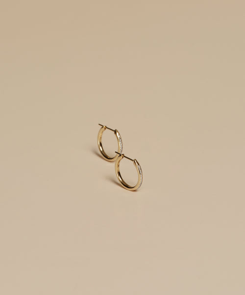 A pair of Iris Diamond Hoops in 14k gold, adorned with subtle diamonds, displayed on a smooth beige surface.