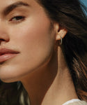 Close-up of a womans face in natural light, showing her clear skin, soft pink lips, and shimmering white gold Iris Diamond Hoops by Jenni Kayne. Her dark hair is gently swept back in front of a softly blurred, blue-tinted background.