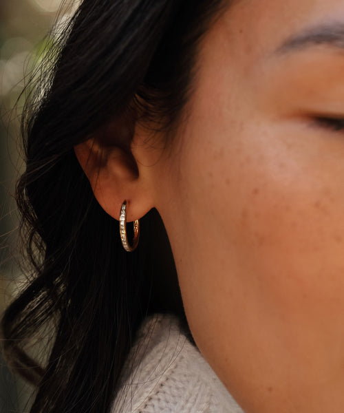 Close-up of a womans ear wearing Jenni Kaynes Iris Diamond Hoops—14k gold earrings with small diamonds. Her eye is closed as her dark hair softly frames her face against a blurred background.