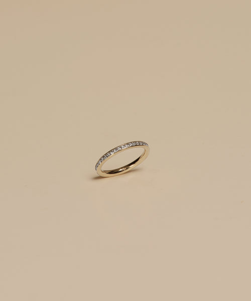 The Iris Eternity Band is a delicate ring crafted in 14k gold, featuring small, evenly spaced conflict-free diamonds along the band, shown against a simple beige backdrop.