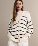 A woman models the Chloe Cardigan in ivory navy stripe with wooden buttons over a white top, hands in white pants pockets, highlighting this stylish sweater against a plain beige background.