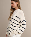 A woman with long brown hair wears the Chloe Cardigan in Ivory Navy Stripe over a white top, standing in profile against a beige background.
