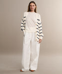 A woman stands against a neutral background wearing the Chloe Cardigan in Ivory Navy Stripe over a white top, paired with wide-leg white trousers and brown shoes. Her hands are in her pockets, offering a chic warm-weather sweater look.