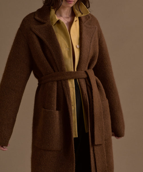 A woman wears the Jessica Sweater Coat, a brown alpaca blend long coat, over a yellow button-up shirt, standing against a plain beige background. Her face is partially out of frame.
