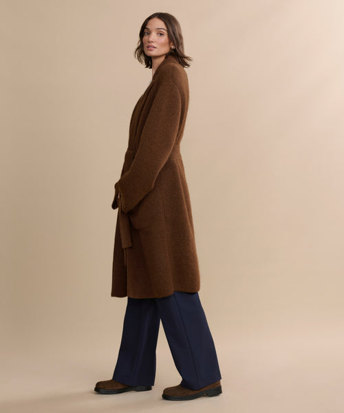 A woman stands sideways in the Jessica Sweater Coat—a long brown alpaca blend with a tie belt—paired with navy blue pants and brown shoes, set against a plain beige background.