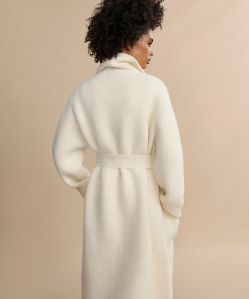 A woman with curly hair is seen from behind, wearing the Jessica Sweater Coat—an ivory, long alpaca blend style with a high collar and matching belt—against a plain beige background.