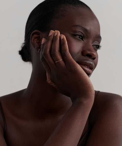 A woman with dark skin, in a thin strap top, thoughtfully rests her face on her hand. She wears the June Baguette Band, a simple 14k gold ruby ring, her calm expression standing out against a neutral background.