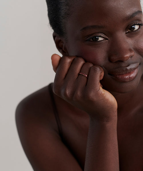 A woman with dark skin smiles softly, resting her chin on her hand. She wears the June Baguette Band on her finger and a thin strap top, set against a plain, light background.
