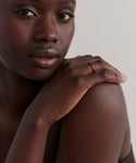 A close-up of a woman with dark skin resting her hand on her bare shoulder, highlighting the June Baguette Band—a thin 14k gold ring—on her finger as she gazes softly at the camera against a plain background.