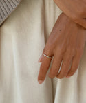 A close-up of a hand wearing the June Baguette Band, a simple 14k gold ring, on the ring finger. The hand rests against cream fabric with part of a beige sweater sleeve visible in the top left.