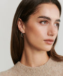 A woman with straight brown hair wears a beige fuzzy sweater and the June Baguette Hoops in white gold, gazing calmly to the side against a light background.