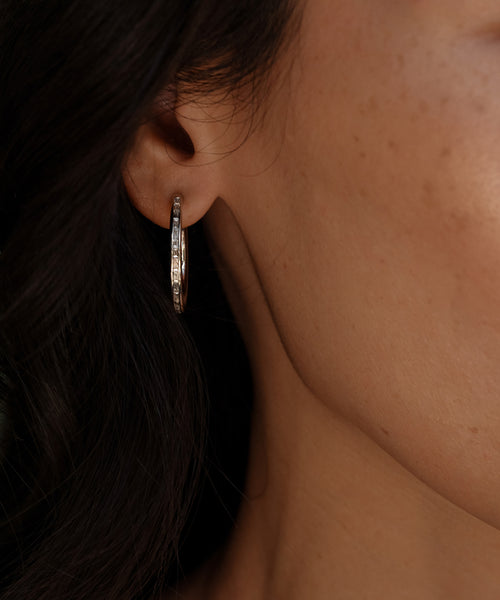 Close-up of a womans ear wearing Jenni Kaynes June Baguette Hoops—a gold hoop earring with a textured design. Her clear, freckled skin and dark hair are partially visible along the edge of the image.