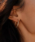 A close-up of a persons ear wearing June Baguette Hoops in 14k gold with a row of sparkling stones. Their dark hair frames the scene, and smooth, evenly lit skin highlights the elegant detail of these diamond earrings.