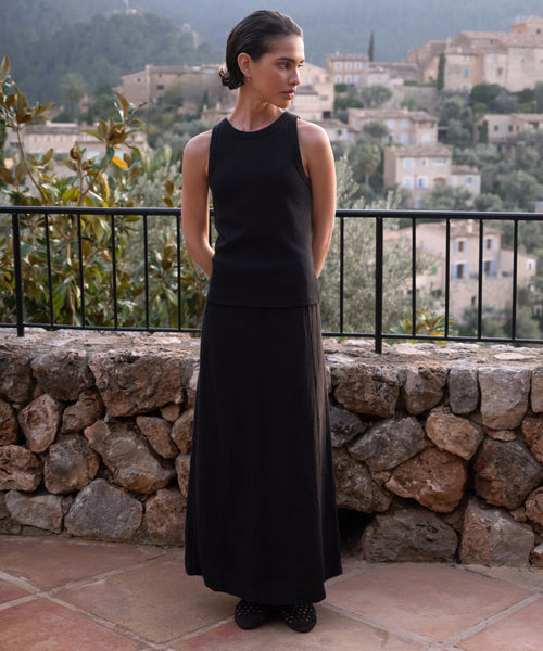 A woman with short, dark hair in a sleeveless black top and the Kinney Skirt stands on a stone patio, hands behind her back. Behind her is a stone wall and a picturesque hillside village.