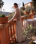 A woman in a striped sweater and the Kinney Skirt stands on a terrace with potted plants, glancing over her shoulder. Behind her, a sunlit table set for dining and lush greenery complete the scenic backdrop.