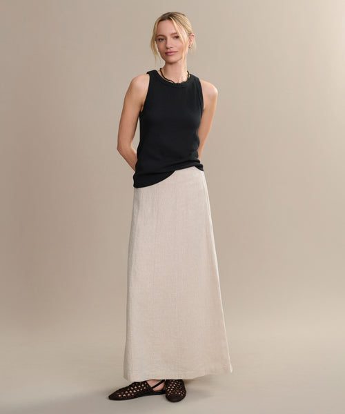 Against a plain beige background, a woman wears the Kinney Skirt—a long, lightweight linen skirt—paired with a sleeveless black top and black shoes. Her arms are relaxed behind her back as she gazes gently to the side.
